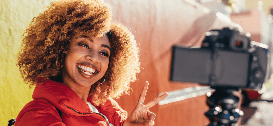 travel write-offs (1) A cheerful woman with curly blonde hair flashes a peace sign at a DSLR camera on a tripod while traveling in a red jacket against a warm-toned wall.